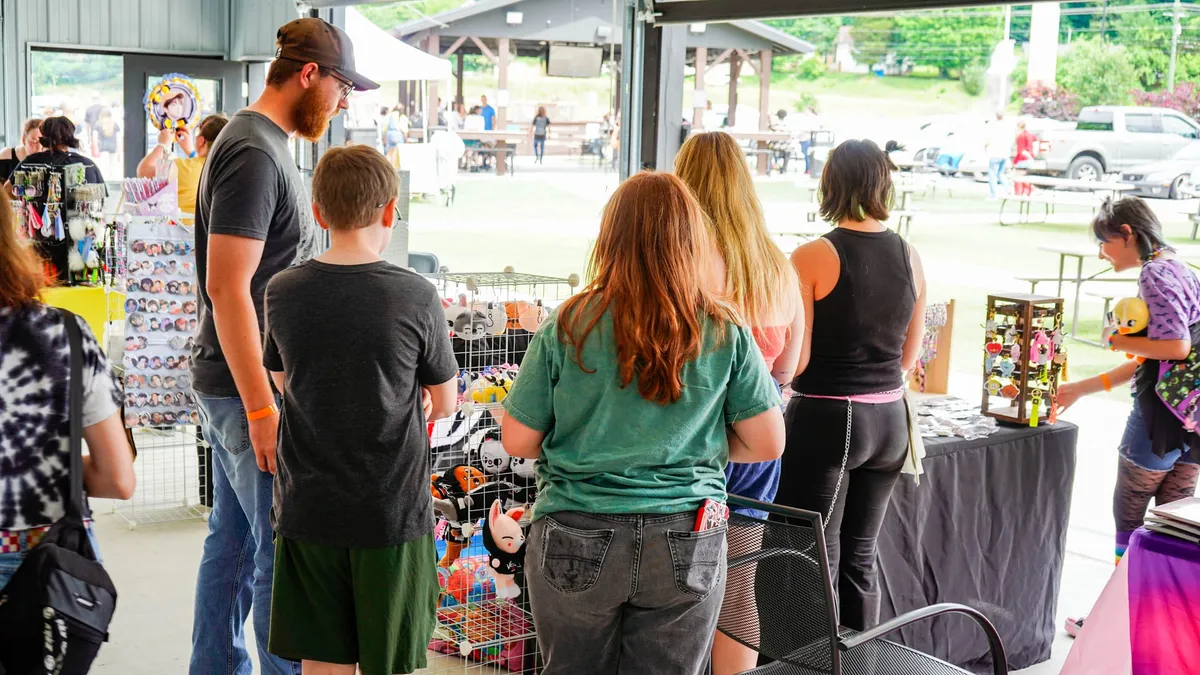 Shoppers including a man and a young boy look over spinning racks of keychains and baskets of plushies at merch tables in an open-air covered pavilion at Knox Pop Con.